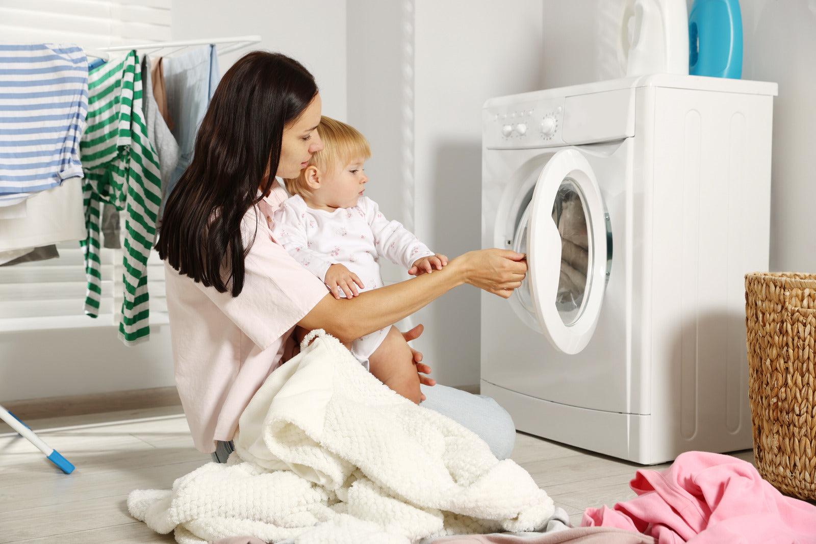 Woman holding a baby and opening a washing machine in a laundry room.