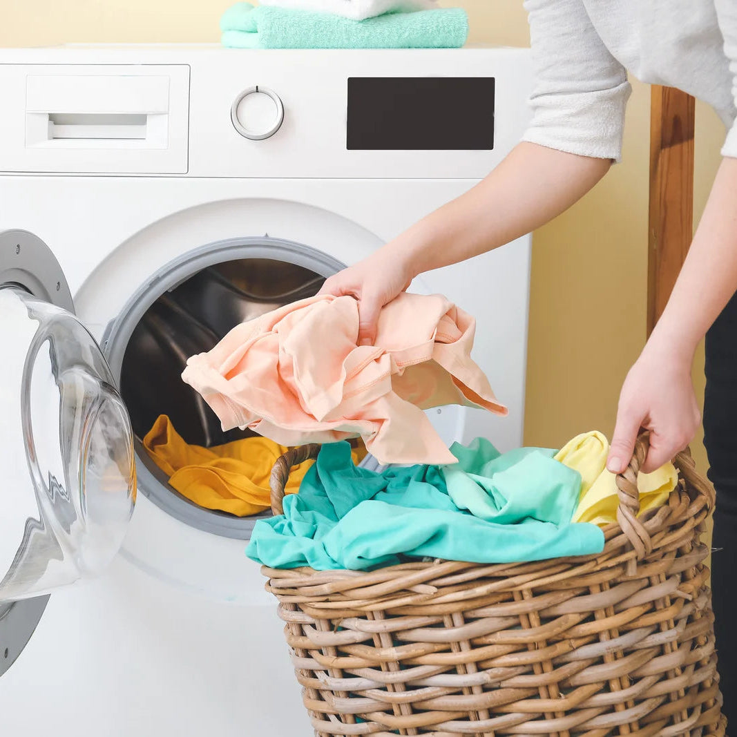 Person loading clothes into a washing machine with a basket of laundry nearby.