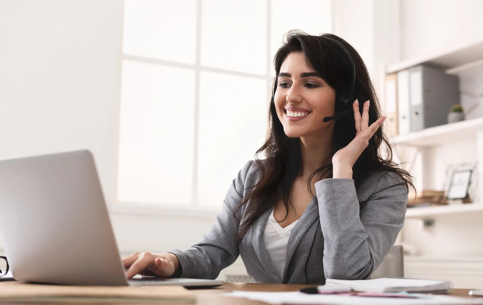 Woman with a headset smiling and using a laptop in a bright room.